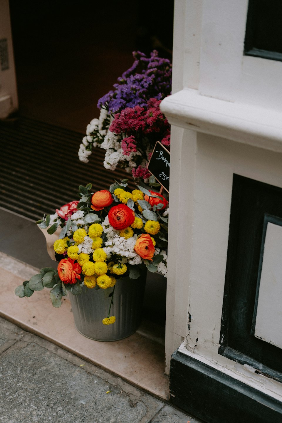 Orange ranunkler och gula blommor i en grå hink på ett stengång.
